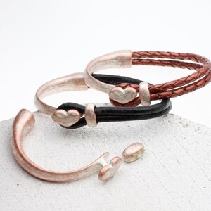 May include: Three bracelets with heart-shaped clasps. One is a silver-tone cuff, and the other two have leather straps in black and brown. The bracelets are displayed on a white surface.