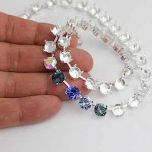 Puede incluir: Tres hebras de cadena plateada con engastes de garra para diamantes de imitación. La cadena se muestra con tres diamantes de imitación de diferentes colores: iridiscente, azul y azul oscuro.