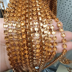 Puede incluir: Primer plano de múltiples hebras de cadena de pedrería dorada. Las cadenas están densamente empaquetadas, mostrando el intrincado diseño de los engastes individuales de pedrería. Las cadenas se sostienen en una mano, con el fondo desenfocado.