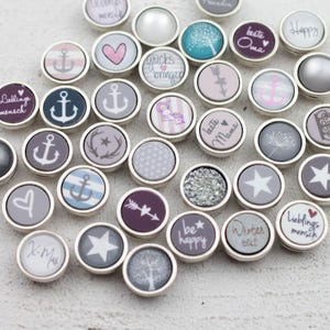 Könnte beinhalten: Eine Sammlung runder Metallknöpfe mit verschiedenen Motiven wie Ankern, Herzen, Sternen, Bäumen und Texten wie "beste Mama", "glucks bringer" und "Lieblingsmensch". Die Knöpfe weisen vielfältige Muster in Grau-, Blau-, Rosa- und Weißtönen auf.