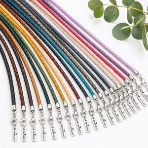 Pode incluir: Uma cole&ccedil;&atilde;o de cord&otilde;es de couro tran&ccedil;ado em v&aacute;rias cores, incluindo preto, castanho, verde, amarelo, azul, cinzento, rosa e roxo. Cada cord&atilde;o possui um fecho lagosta de metal prateado e uma etiqueta gravada com o texto CRISTALOGY.