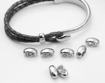 May include: A bracelet featuring a dark braided cord and a polished silver-tone metal hook clasp, accompanied by several small, smooth, oval-shaped silver-tone metal beads with two holes each, arranged in a cluster below the bracelet.