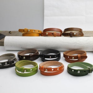 May include: A collection of eleven leather wrap bracelets in various colors including yellow, brown, black, green, and orange. Each bracelet features a thin leather strap, a metal stud closure, and multiple adjustment holes for a secure fit.