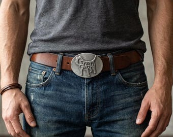 May include: A brown leather belt featuring a silver-toned oval buckle with a raised relief design of a dog and the text Gran Can. The belt is worn with blue denim jeans and a grey t-shirt, with a brown leather wristband on the left arm.