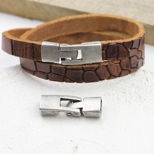Könnte beinhalten: Ein braunes Lederarmband mit einem silberfarbenen Verschluss. Das Armband hat ein krokodilgeprägtes Muster.