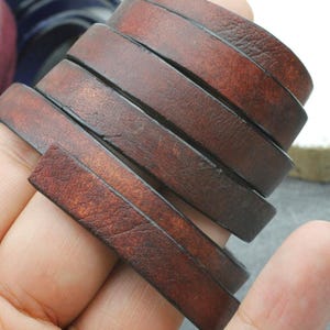 Könnte beinhalten: Ein Stapel von sechs braunen Lederarmbändern. Die Armbänder bestehen aus flachen, rechteckigen Lederstreifen, die jeweils einen leicht unterschiedlichen Braunton aufweisen. Das Leder hat ein strukturiertes, abgenutztes Aussehen.