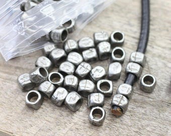 May include: A close-up of a pile of silver metal beads with runes engraved on them. The beads are scattered on a wooden surface. A black leather cord is visible in the background.