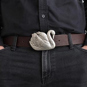 May include: A dark brown leather belt featuring a large, silver-toned metal buckle shaped like a swan. The swan buckle displays detailed feather textures and a curved neck, secured onto a smooth, solid-colored brown leather strap with a standard prong.