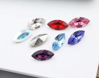 May include: A collection of eight marquise-shaped faceted crystals in various colors including clear, red, pink, iridescent, silver, light blue, dark blue, and purple, arranged in a scattered pattern on a plain white surface.