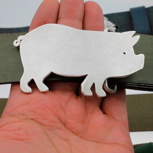 Peut inclure: Une boucle de ceinture en argent en forme de cochon. La boucle est en forme de cochon et a un contour détaillé. La boucle est attachée à une ceinture en cuir vert.