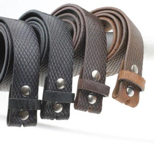 Op de afbeelding: Vier opgerolde riemen met een gestructureerd ruitpatroon. De riemen zijn gerangschikt in een verloop van zwart naar donkerbruin en lichtbruin. Elke riem is voorzien van een leren lus en twee zilverkleurige metalen drukknopen aan het uiteinde.