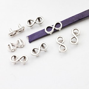 May include: Silver infinity symbol charms with a hole for leather cord. The charms are scattered on a white surface with a purple leather cord in the background.