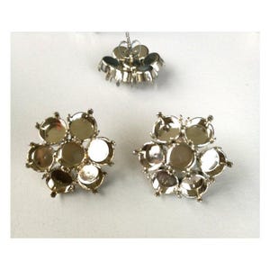 Puede incluir: Tres componentes de pendientes de metal plateado. Dos tienen forma de flor con siete engastes circulares, y uno es un cierre de tipo poste. Los componentes están diseñados para sujetar piedras redondas o adornos para la fabricación de joyas.