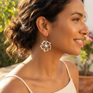 May include: A silver-toned drop earring featuring a stylized five-petaled flower design with an openwork structure and a central circular bead, suspended from a thin metal hook that passes through the earlobe for a secure fit.