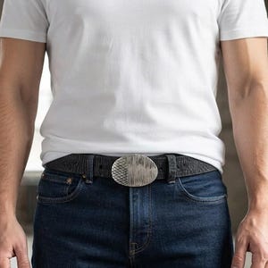 May include: A black leather belt with a textured surface and an oval-shaped silver-toned metal buckle. The buckle features a split design with a cross-hatched grid pattern on the left side and vertical brushed lines on the right side.