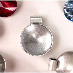 May include: A silver pendant with a circular setting and a loop for a chain. The pendant has a textured surface and is surrounded by other gemstones. The pendant is a jewellery component.