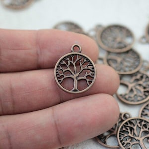 May include: Bronze-colored tree of life charm. The charm is circular with a detailed tree design inside. The charm has a small loop at the top for attaching to jewelry or other crafts. Several similar charms are visible in the background.