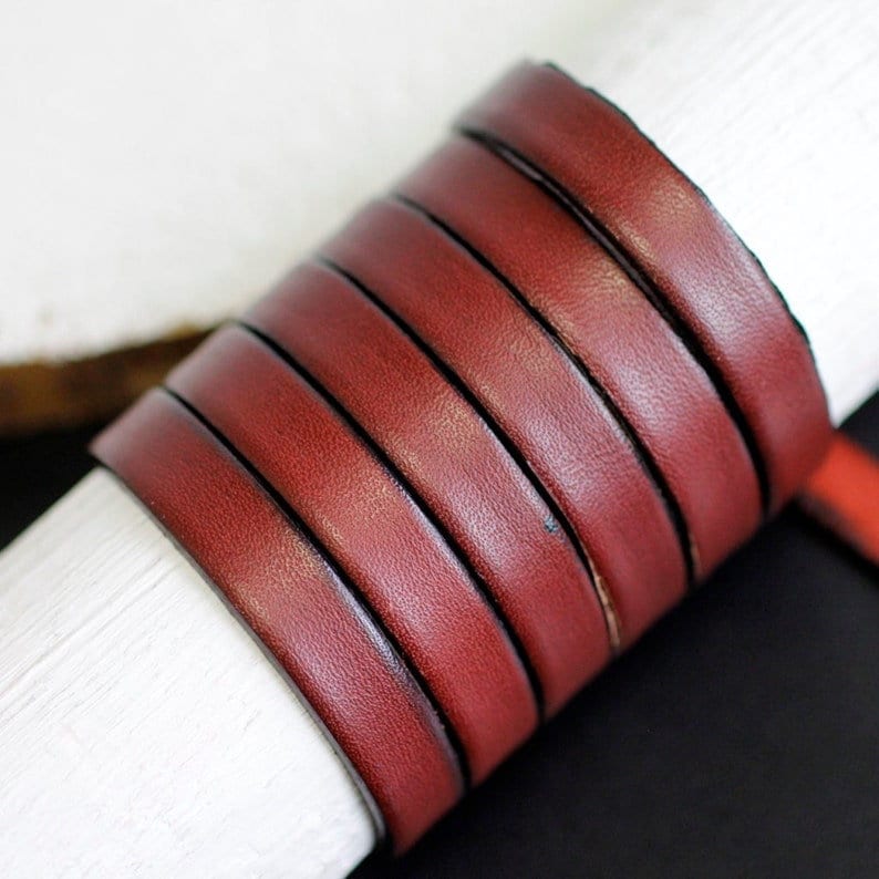 May include: A close-up of a roll of burgundy leather cord. The cord is approximately 5/16 inch wide and has a smooth, polished finish.
