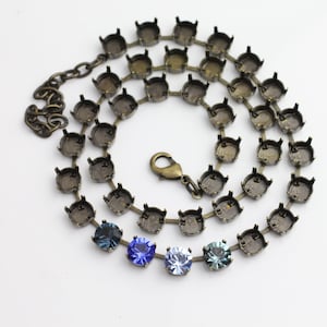 Puede incluir: Un collar con una cadena de color bronce y engastes para piedras preciosas. El collar tiene un cierre y presenta cuatro piedras preciosas de colores: azul oscuro, azul real, azul claro y verde azulado. El collar se muestra sobre una superficie blanca.