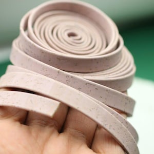 Puede incluir: Un rollo de cinta de corcho rosa claro con textura moteada. La cinta está enrollada y se sostiene en una mano.