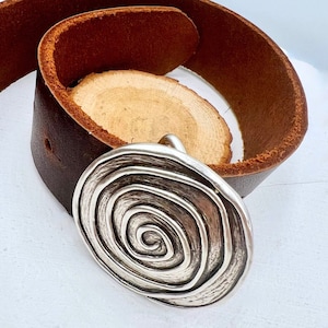 Pode incluir: Um pingente prateado em forma de espiral com um cinto de couro castanho escuro. O pingente tem uma superfície texturizada. O cinto é enrolado em uma fatia de madeira natural.