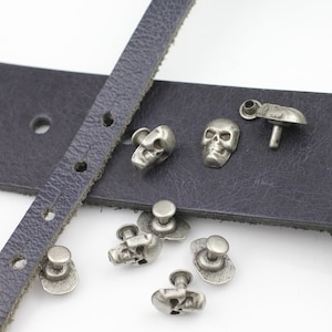 Puede incluir: Primer plano de remaches plateados con forma de calavera y un cinturón de cuero azul oscuro. Los remaches están en varias posiciones, algunos sujetos al cinturón y otros sueltos. Los remaches tienen un diseño detallado de calavera.