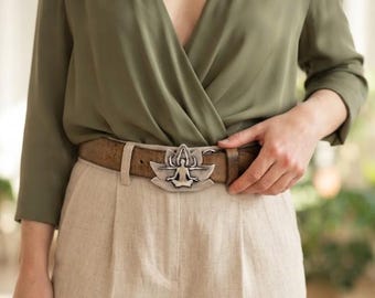 May include: A brown leather belt featuring a silver-toned metal buckle shaped like a lotus flower with a central figure in a seated meditation pose. The belt is worn with a solid olive green long-sleeved blouse and light beige high-waisted trousers.