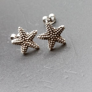 Puede incluir: Un par de pendientes de estrella de mar de plata. Los pendientes están hechos de pequeñas cuentas redondas que se agrupan para formar la forma de una estrella de mar.