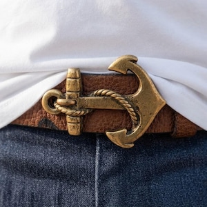 May include: A brown textured leather belt featuring a large, antique brass-toned metal buckle shaped like a nautical anchor with a rope detail wrapped around the center shank, worn over dark blue denim jeans and a plain white t-shirt.
