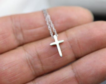 May include: A small, silver-colored cross pendant necklace. The cross is simple in design and hangs from a delicate chain. The necklace is shown against a blurred background.