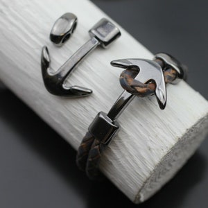 Pode incluir: Duas pulseiras de couro com pingentes de âncora prateados. Uma pulseira é de couro marrom com um design trançado e a outra é de couro preto com um design simples.