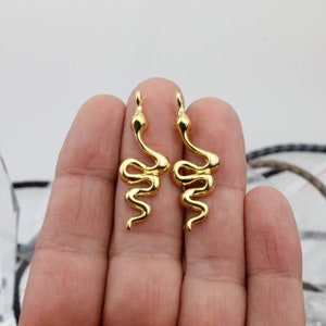 Puede incluir: Dos colgantes de serpiente de color dorado. Los colgantes de serpiente están enrollados y tienen un acabado liso y brillante.