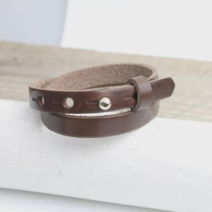 Peut inclure: Un bracelet en cuir marron foncé avec des rivets argentés et une boucle. Le bracelet s'enroule deux fois, mettant en valeur son design. Le cuir a une texture naturelle et une riche couleur marron foncé. Environ 20 cm de long.