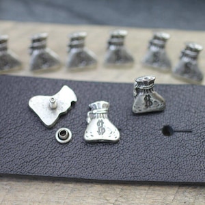 May include: Silver-toned metal bag-shaped studs with a dollar sign, along with a matching backing piece and a small washer. The studs are arranged on a black leather surface, suggesting a design element for leather goods or accessories.