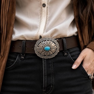 Op de afbeelding: Een donkerbruine leren riem met een grote, ovale gesp van zilverkleurig metaal. De gesp heeft een reliëf van bloemblaadjes met in het midden een gladde, ovale steen in een turquoise kleur, gedragen over een zwarte spijkerbroek.