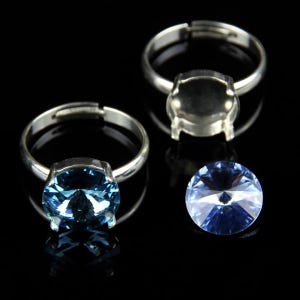 May include: Three silver adjustable rings with a round, light blue gemstone. One loose, faceted, light blue gemstone is also present. The rings and gemstone are set against a black background.