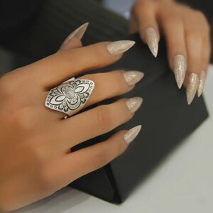Könnte beinhalten: Ein silberfarbener Ring mit einer länglichen, schildförmigen Vorderseite, die mit komplizierten schwarzen, gravierten Blumen- und Geometriemustern versehen ist. Der Ring wird am Ringfinger getragen und zeigt ein detailliertes, symmetrisches Design.