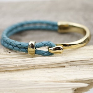May include: A braided blue leather bracelet with a gold metal clasp. The bracelet is on a light wood surface.