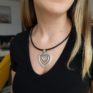 Könnte beinhalten: Halskette mit silbernem Herzanhänger. Der Anhänger hat drei ineinander verschachtelte Herzen, die an einem schwarzen Band hängen. Die Halskette wird über einem schwarzen V-Ausschnitt-Shirt getragen.