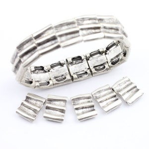May include: A silver bracelet with a textured design. The bracelet is made of multiple linked pieces and has a clasp closure. There are five extra pieces of the bracelet lying on a white surface.
