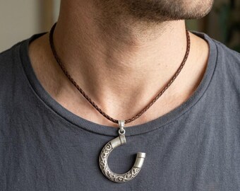 Può includere: Un ciondolo in metallo color argento a forma di ferro di cavallo stilizzato con intricate incisioni a volute. Il ciondolo pende da un sottile cordino in pelle intrecciata marrone scuro che poggia sul collo, con un terminale metallico liscio.