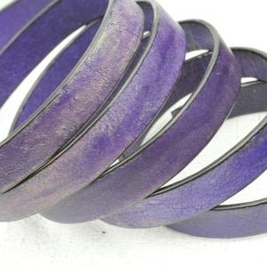 Puede incluir: Primer plano de un cinturón de cuero morado enrollado. El cinturón tiene una superficie texturizada con un ligero brillo. Los bordes son oscuros y el cuero parece ligeramente desgastado, dándole un aspecto vintage. El fondo es blanco.