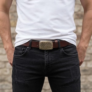 May include: A brown leather belt featuring a rectangular brass buckle with a textured, hammered finish. The belt is worn through the loops of black denim jeans, secured with a single prong, and includes a matching leather keeper loop.