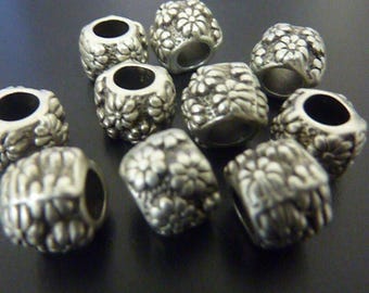 May include: Close-up of several silver-colored beads with a floral design. Each bead has a cylindrical shape with a large hole running through the center. The beads are clustered together on a dark surface.
