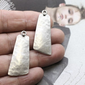 May include: Two silver-toned metal pendant charms with a hammered texture. The charms are rectangular with rounded corners and have a small loop at the top for attaching to a chain or cord.