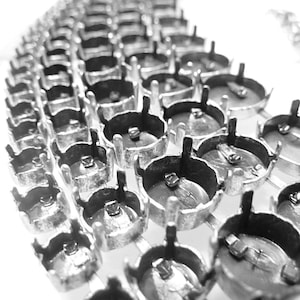 Puede incluir: Primer plano de una colección de engastes de joyería de metal plateado. Los engastes están dispuestos en filas, cada uno con una base circular y garras que se extienden hacia arriba. La imagen es en blanco y negro, resaltando el brillo metálico.