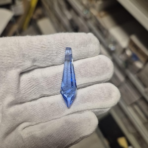 May include: A blue crystal pendant with a pointed end and a rectangular top, held in a gloved hand. The pendant's facets are visible, reflecting light. The crystal's design and colour are highlighted, showcasing its intricate details. The pendant is a striking piece.