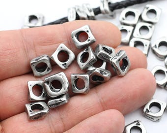 May include: A collection of small, square-shaped metal beads with a distressed silver finish. Each bead features a central circular hole and a textured, uneven surface. Some beads are loose, while others are threaded onto a black braided cord.
