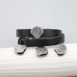 Könnte beinhalten: Ein schwarzes Lederarmband mit silberfarbenen Muschel-Anhängern. Das Armband ist zweimal um sich selbst gewickelt und die Anhänger sind auf beiden Seiten sichtbar.