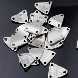 May include: A pile of silver triangle-shaped metal charms with three holes each. The charms have a textured surface and are ideal for jewellery making.